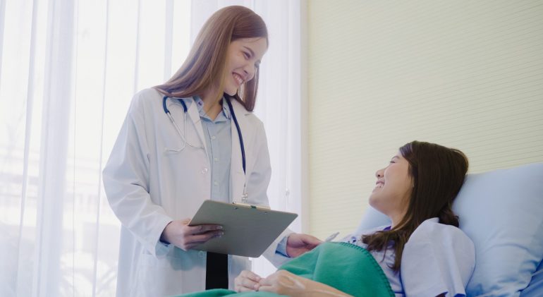Beautiful smart Asian doctor and patient discussing and explaini Beautiful smart Asian doctor and patient discussing and explaining something with clipboard in doctor hands while staying on Patient's bed at hospital. Medicine and health care concept.